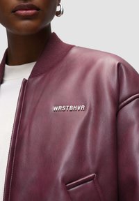 Burgundská kožená bomber bunda má žebrovaný límec a stříbrné vyšívané logo "WRSTBHVR" na přední straně. Hladká textura, zapínání na zip.