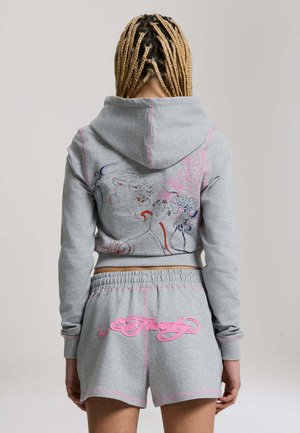 Person med fletninger iført et gråt hættetrøje- og shortsæt med pink syning, broderet tiger- og blomsterdesign på ryggen.