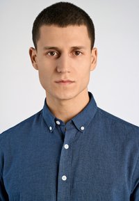 Jonge man met kort bruin haar en bruine ogen, gekleed in een lichtblauw overhemd met knopen, kijkt recht vooruit tegen een effen lichte achtergrond.