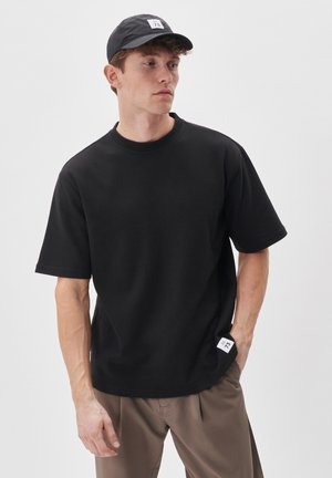 Czarny t-shirt z krótkim rękawem z okrągłym dekoltem, wykonany z bawełny. Posiada boczną metkę z logo marki. Noszony z pasującą czarną czapką.