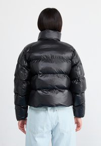 Sort pufferjakke med høj krave, der har quiltede sektioner, blank overflade og cropped design. Båret over lyseblå jeans.
