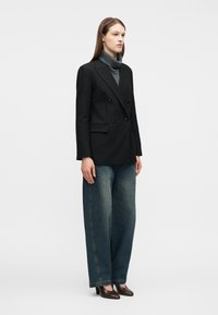 Γυναίκα που στέκεται φορώντας μαύρο διπλό κουμπωτό σακάκι, γκρι πουλόβερ με ψηλό λαιμό, ανοιχτόχρωμο τζιν με φαρδιά πόδια και καφέ υφασμάτινες ψηλοτάκουνες γόβες.