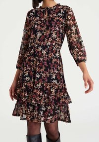 Robe noire à fleurs avec de longues manches transparentes, un ourlet volé et une taille élastique. Présente de petits motifs floraux dans des tons beige et rose.
