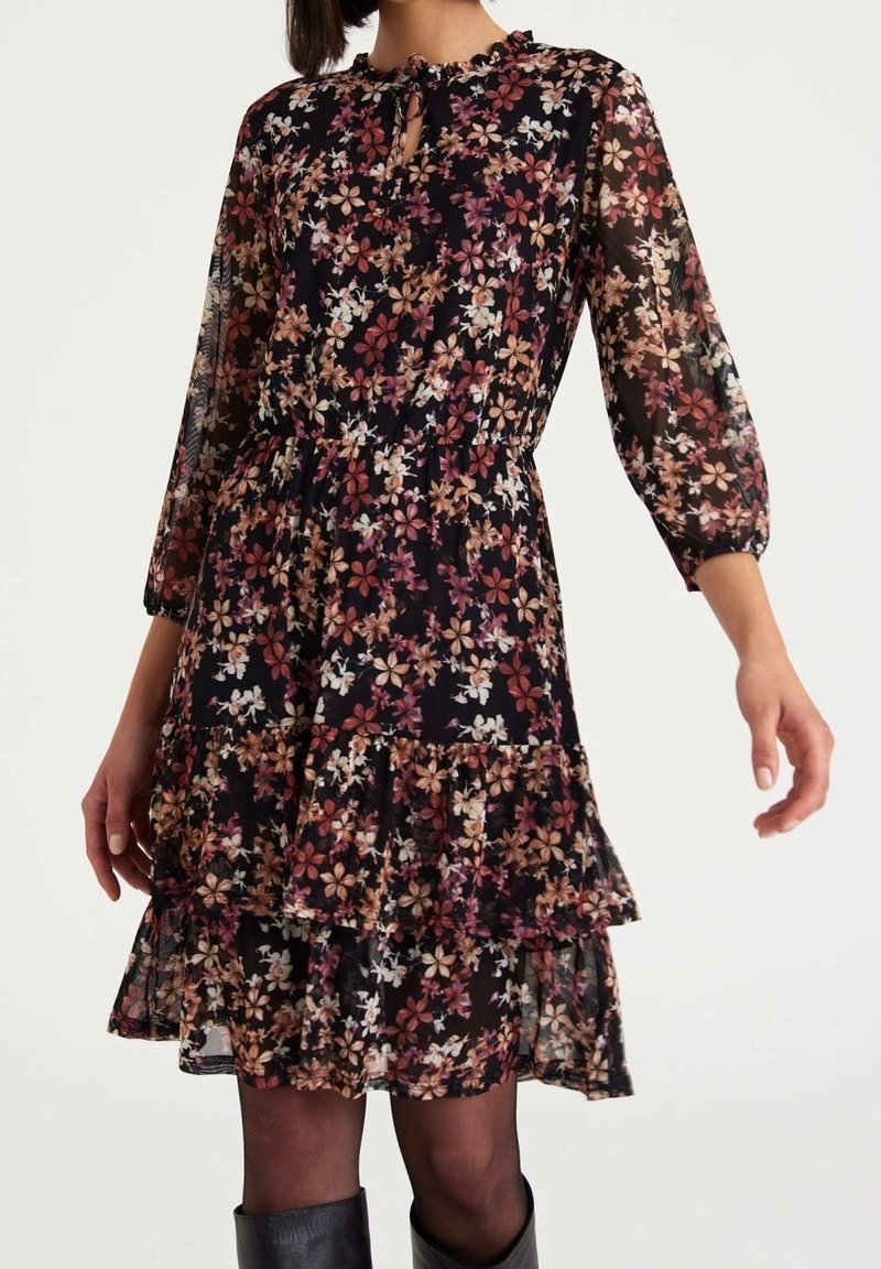 Robe noire à fleurs avec de longues manches transparentes, un ourlet volé et une taille élastique. Présente de petits motifs floraux dans des tons beige et rose.