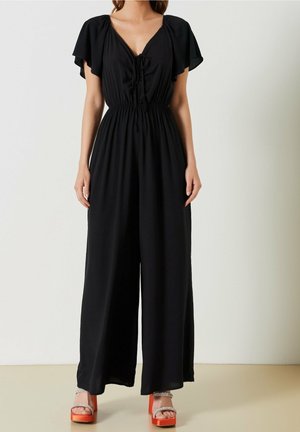 Kobieta ubrana w czarny kombinezon z szerokimi nogawkami, z falującymi rękawami i dekoltem w kształcie litery V, zestawiony z pomarańczowymi sandałami na platformie z paseczkami ozdobionymi kryształkami.