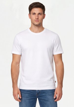 Giovane uomo in piedi rivolto verso il fronte, indossa una t-shirt bianca a maniche corte e un paio di jeans blu su uno sfondo bianco.