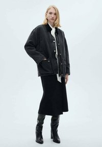 Black denim jacket with snap buttons, front pockets, layered over a white blouse and black skirt. Knee-high black boots complete the outfit.