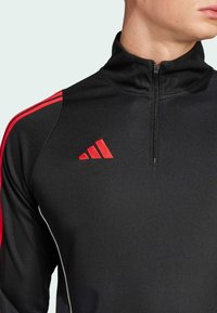 Hombre vistiendo una camiseta deportiva negra de media cremallera con rayas rojas en las mangas y el logotipo rojo de Adidas en el pecho, mostrado desde el cuello hasta la mitad del torso.