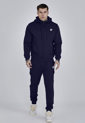 Mann steht, trägt einen marineblauen Kapuzenpullover mit Reißverschluss und passende Cargo-Jogginghosen sowie weiße Turnschuhe vor einem einfarbigen grauen Hintergrund.