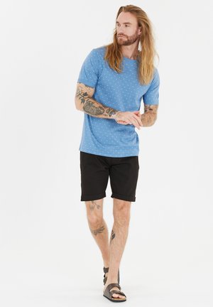 Chemise bleue à manches courtes avec des pois blancs, associée à un short noir et des sandales marron. La chemise présente un col rond et une coupe décontractée.