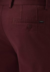 Burgundowe materiałowe spodnie z tylną kieszenią na guzik i małą tkana metką. Tekstura jest gładka z delikatnym połyskiem.