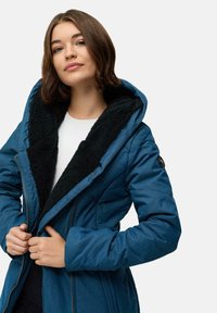 Ung kvinne som drar glidelåsen på en blå vinterjakke med svart fleecefôr og hette, iført hvit skjorte under, nøytral bakgrunn.