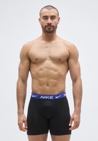Boxeri negri cu talie albastră cu logo-ul Nike, cu o croială strâmtă și textură netedă, purtați de un model masculin fără tricou.