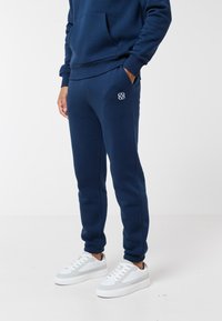 Asmuo dėvi tamsiai mėlynas sportines kelnes su siuvinėta logo ant šlaunies, derinančias su tokio paties stiliaus džemperiu su kapišonu ir baltus sportbačius, stovėdamas ant baltos grindų.