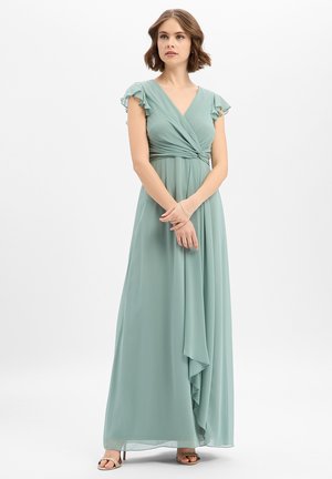Vrouw staand gekleed in een lange wikkeljurk in saliegroen met vleermuismouwen en open sandaaltjes tegen een effen witte achtergrond.