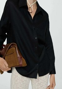 Camicia nera con colletto, maniche lunghe e texture liscia. Tiene una borsa marrone e rossa. Presenta un dettaglio unico a nodo sul collo.