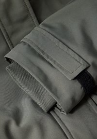 Polsino di giacca verde oliva con una finitura liscia e opaca, foderato in maglia a coste e dotato di una fascia di regolazione con Velcro. Le cuciture sono visibili.