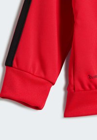 Camiseta deportiva roja de manga larga con puños acanalados, tejido texturizado y acentos laterales negros. Incluye una etiqueta "CLIMACHILL" en el lado inferior.