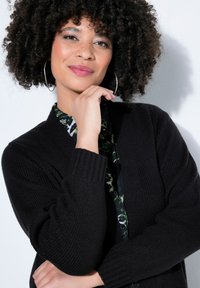 Femme aux cheveux bouclés, portant des boucles d'oreilles créoles et une chemise à motif floral sombre sous un cardigan noir, reposant le menton sur la main et souriant doucement devant un fond blanc.