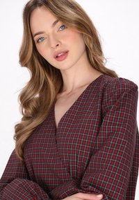 Robe portefeuille en tartan rouge et noir avec un décolleté en V profond, des manches longues et des détails en tissu froncé. Texture douce avec un motif à carreaux.