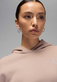 Bluza z kapturem w miękkim, jasnoróżowym materiale z białym logo Jumpman. Modelka ma kolczyki w kształcie kółek i kolczyk w nosie. Gładka tekstura.