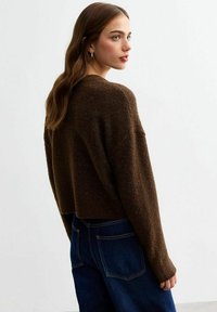 Maglione corto marrone con una finitura testurizzata e maniche lunghe, abbinato a jeans di denim blu scuro. La vista dal retro mette in risalto la vestibilità comoda.