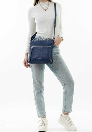 Borsa a tracolla blu in materiale testurizzato, con una tasca frontale con zip e una tracolla regolabile. Indossata con jeans chiari e sneakers.