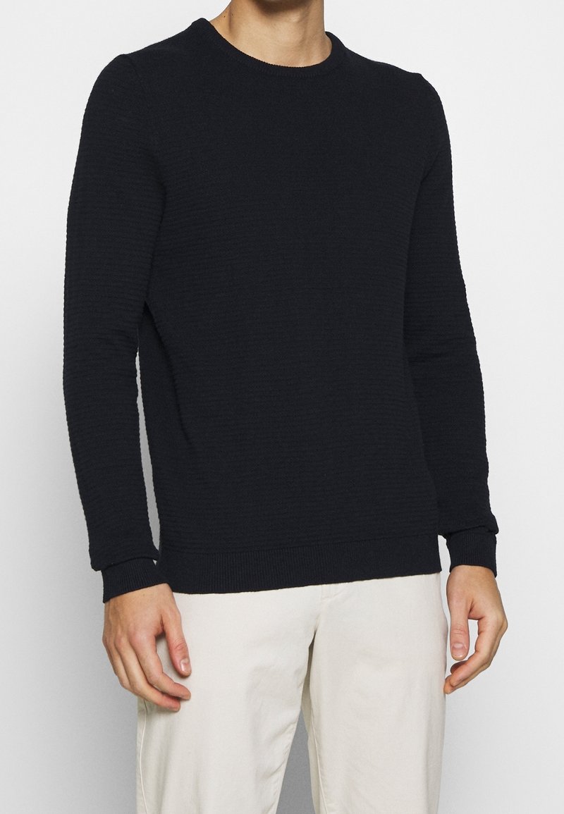 Homme portant un pull noir texturé à manches longues et un pantalon beige clair, debout devant un fond blanc uni.