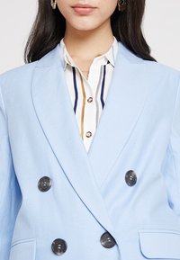 Světle modrý dvouřadý blejzr s klopami s výřezy, černými knoflíky a podšitým vnitřkem, doplněný bílou košilí s vertikálními pruhy.