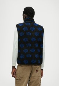 Fekete fleece mellény kék virágmintával, magas gallérral és texturált felülettel. Világos színű hosszú ujjú ing felett viselve.