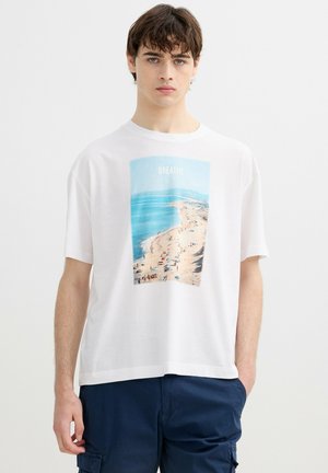 Joven con pelo oscuro que lleva una camiseta blanca con una escena de playa y la palabra "BREATHE", combinada con pantalones azul oscuro.