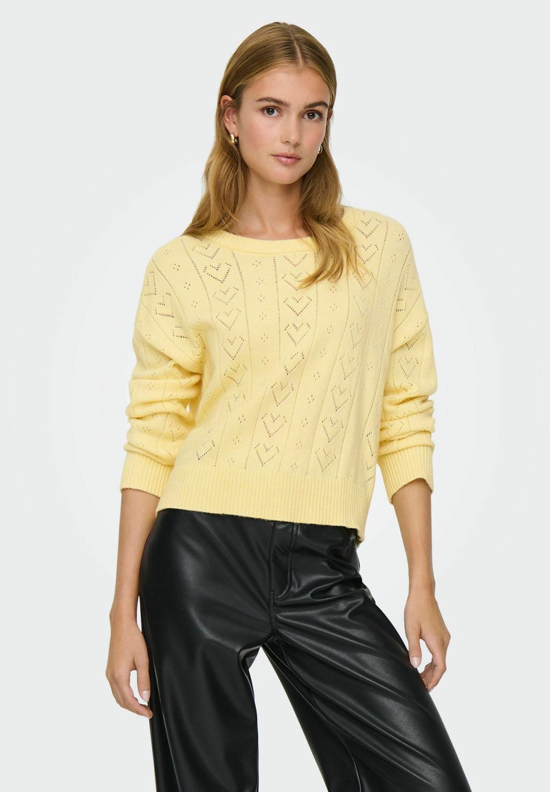 Pull en maille jaune avec un motif losange et cœur, poignets et ourlet côtelés. Associé à un pantalon en cuir noir.