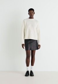 Weißer Strickpullover mit gerippten Bündchen, kombiniert mit einem schwarzen Leder-Minirock. Das Model trägt weiße Socken und klobige schwarze Schuhe.