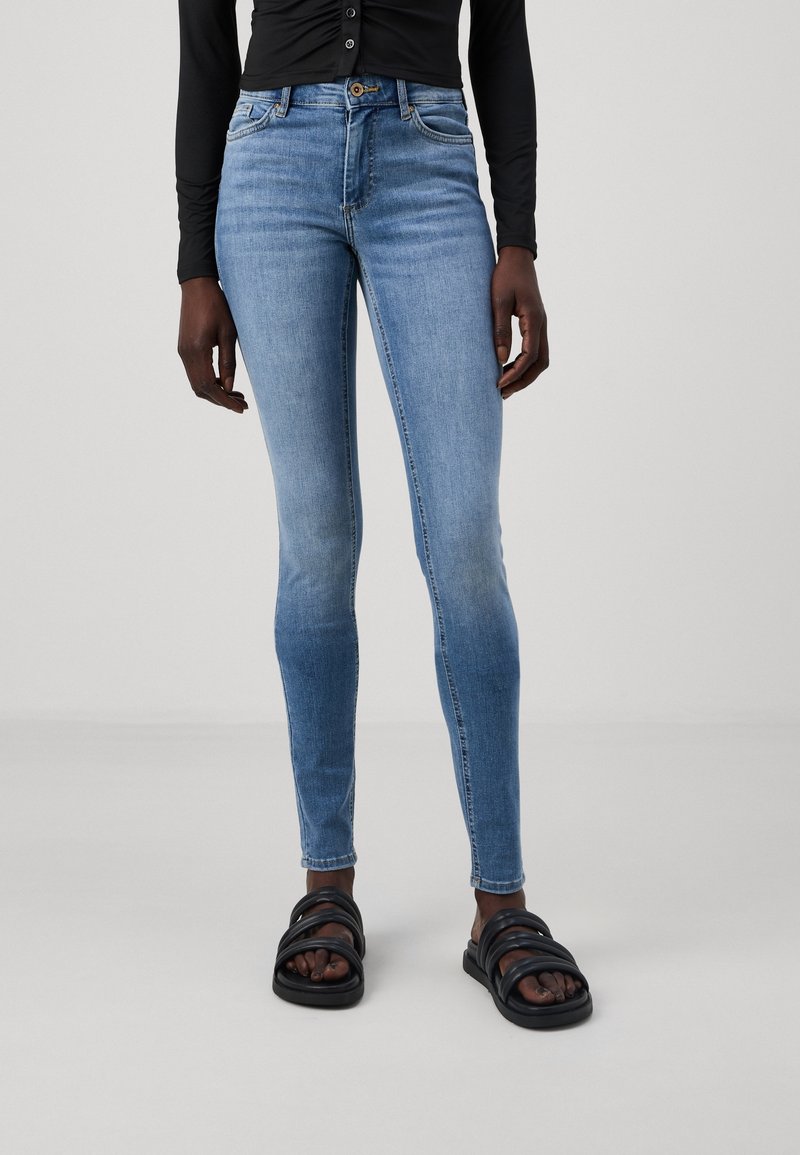 Person i ljusblå smala jeans, svart långärmad topp och svarta vadderade slidesandaler står mot en enfärgad bakgrund.