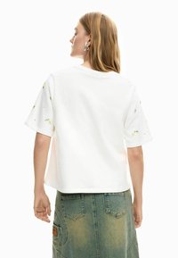 Mujer con el cabello largo, lleva pendientes grandes plateados, una camisa blanca de manga corta con bordados florales y una falda vaquera desgastada, vista desde atrás.