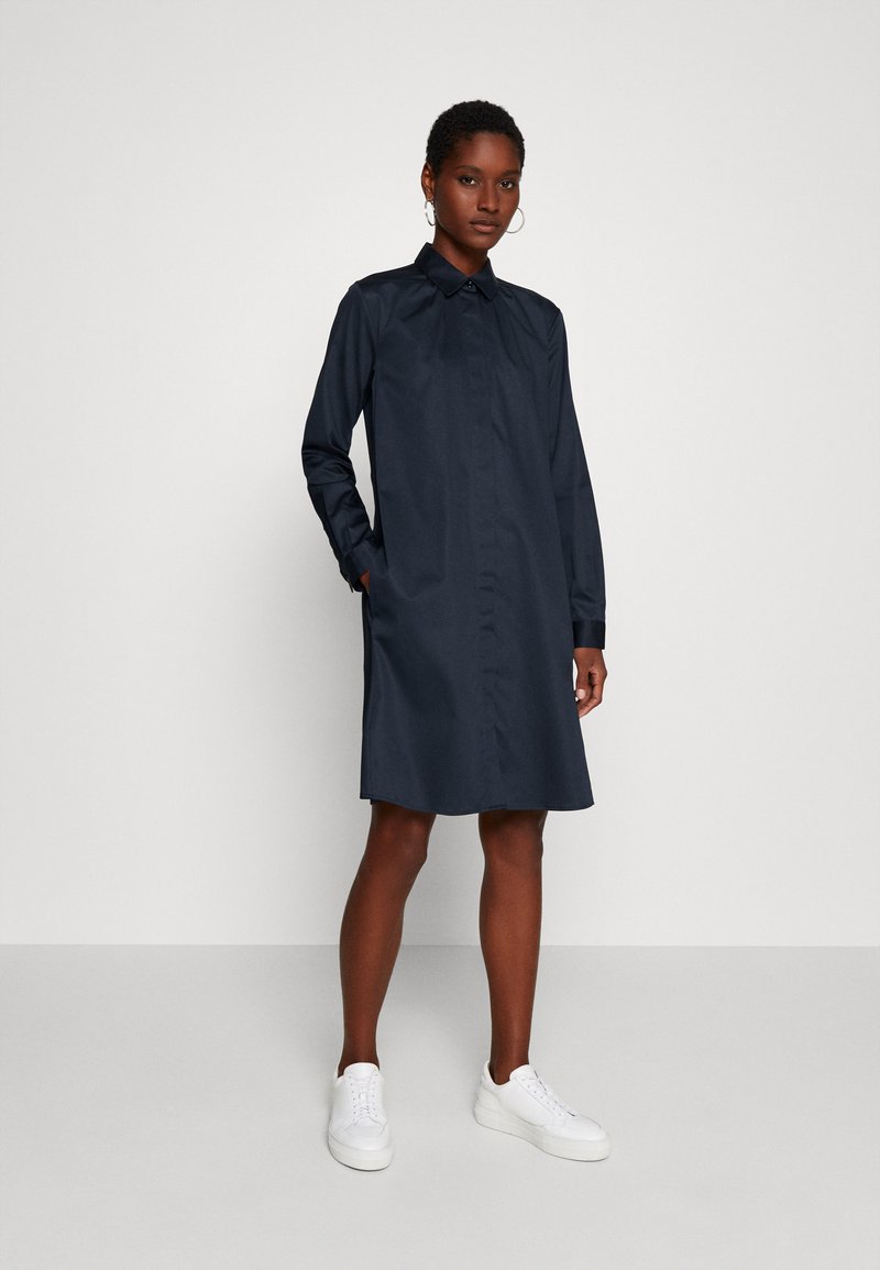 Marineblauwe jurk met een kraag, lange mouwen en zijzakken. Gemaakt van soepele stof, valt boven de knieën en is gecombineerd met witte sneakers.