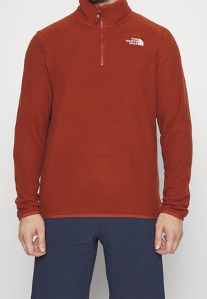 Czerwony polar z ćwiartkowym suwakiem i długimi rękawami, z logo na lewej piersi i gładką fakturą. Połączony z ciemnoniebieskimi spodniami.