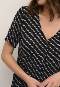 Schwarzes Kleid mit weißem Muster, kurzen Ärmeln, V-Ausschnitt und drei dekorativen goldenen Knöpfen am Oberteil. Weicher, leichter Stoff.