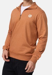 Homme portant un pull à demi-fermeture éclair couleur rouille sur une chemise blanche et un pantalon noir, debout devant un fond uni.