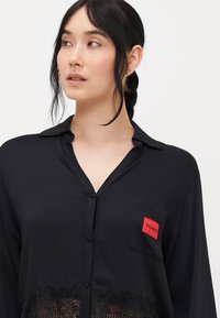 Black button-up shirt with a collar, featuring a lace hem and a red “HUGO” label on the chest pocket. Smooth fabric texture.