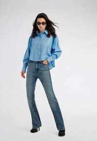 Camicia button-up azzurro chiaro in satin con maniche lunghe, infilata in jeans svasati blu sbiaditi, abbinata a stivaletti neri con tacco e occhiali da sole.