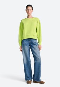 Jasnozielona bluza z okrągłym dekoltem i luźnym krojem, w zestawieniu z szerokimi, niebieskimi jeansami oraz brązowymi butami na sznurowadła.