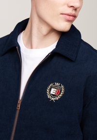 Giacca di velluto a coste blu navy con chiusura a zip, colletto classico e logo ricamato con una corona e alloro, su uno sfondo semplice.