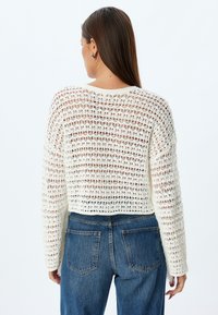 Sweater en crochet blanc avec un design ample et court, à manches longues. Porté par-dessus un jean en denim taille haute, mettant en valeur un style décontracté.