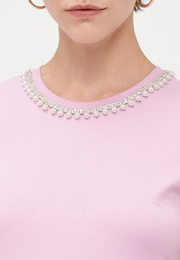 Růžový top s krátkými rukávy, který má kulatý výstřih zdobený řetězy z umělých perel a rhinestones, vytvářejícími texturovanou ozdobu.