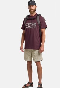 Bordeauxrotes T-Shirt mit grafischem Design, khakifarbene Shorts und schwarze Sandalen. Kombiniert mit einem grauen Rucksack und einer dunklen Mütze. Freizeitliche Outdoor-Bekleidung.
