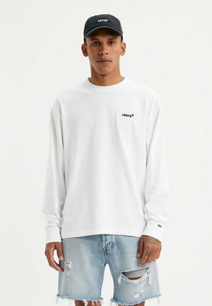 Joven con una camisa blanca de manga larga de Levi's, pantalones cortos de mezclilla azul claro rasgados y una gorra negra de Levi's, de pie contra un fondo liso.