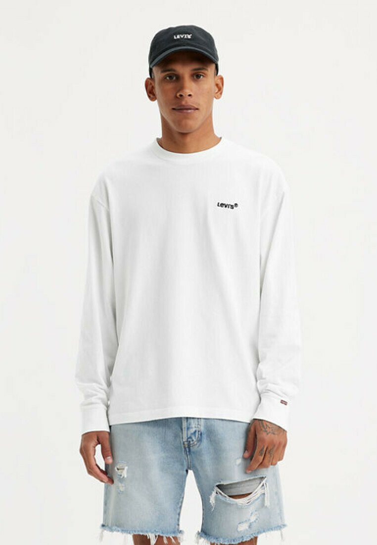 Giovane uomo che indossa una camicia bianca a maniche lunghe Levi's, pantaloncini di jeans azzurri strappati e un berretto nero Levi's, in piedi davanti a uno sfondo semplice.