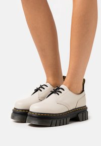 Beige lær snøresko med tykk sort gummisåle, som har gule sting og et teksturert, rillede design.