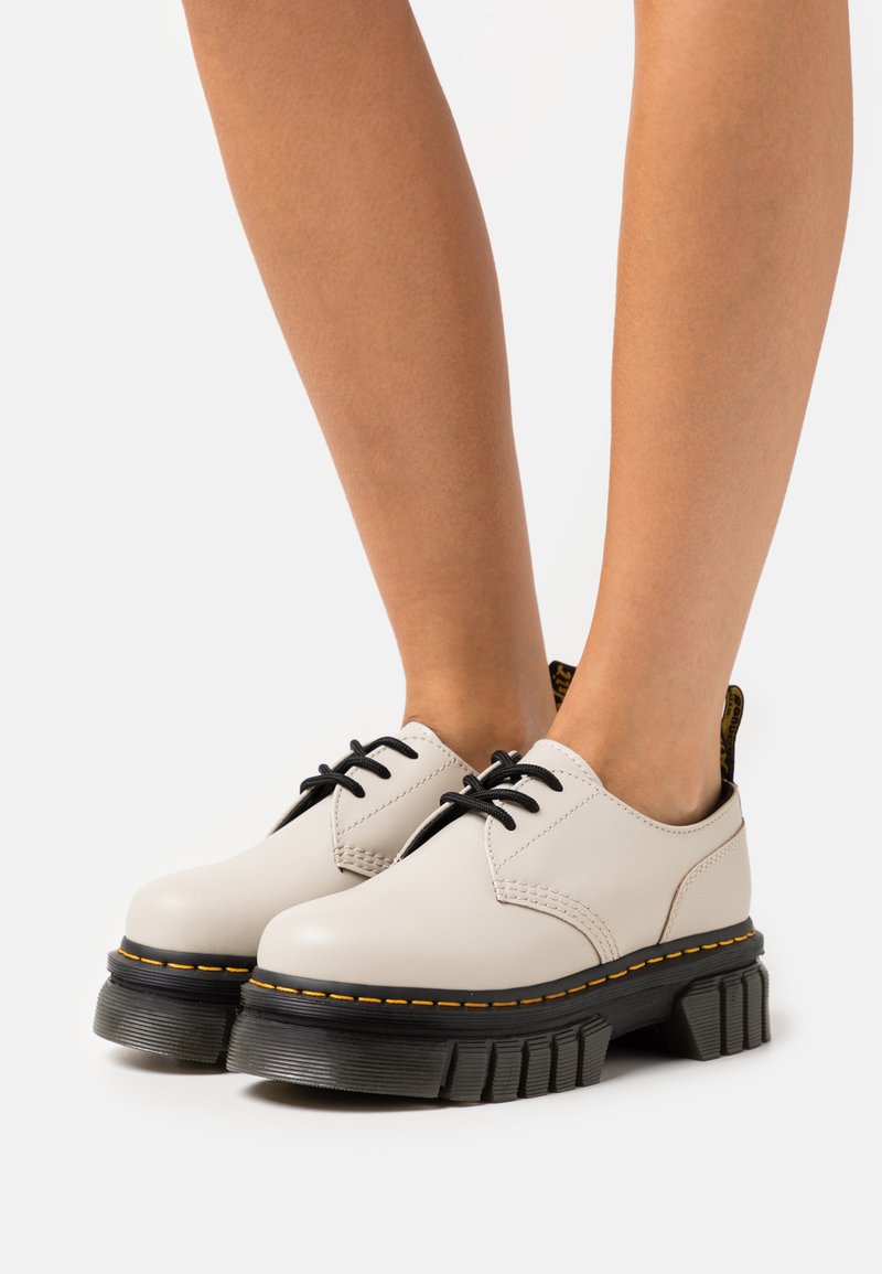 Beige lær snøresko med tykk sort gummisåle, som har gule sting og et teksturert, rillede design.